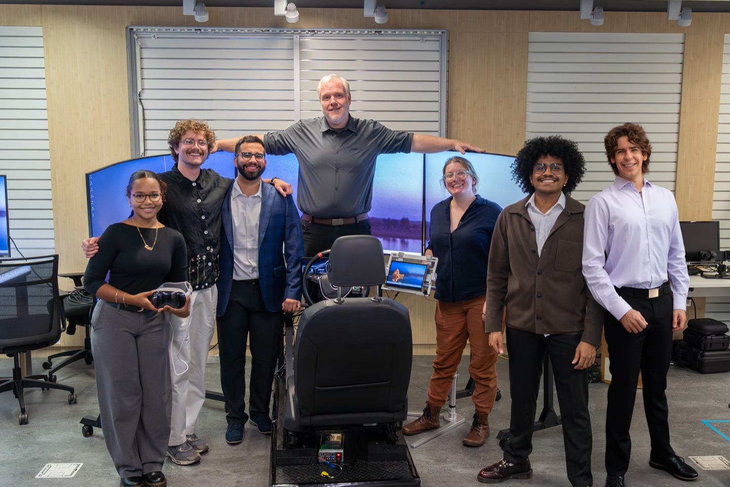Seven members of the lab stand around the driving simulator: its seat, wheel and three curved screens.